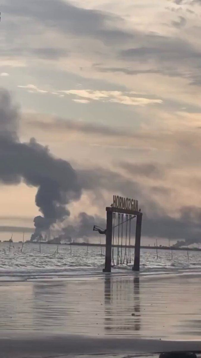 @Visegrád 24: Iranian girl on a swing against a background of smoke rising over Bandar Abbas from a naval base hit by U.S.-Israeli airstrikes